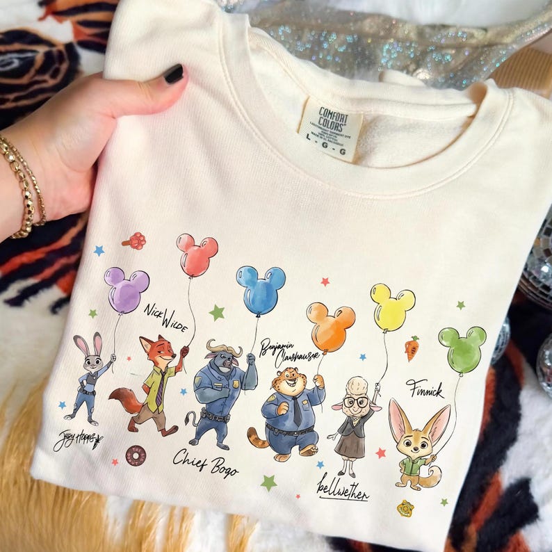 Op de afbeelding: Cr&egrave;mekleurige sweatshirt met stripfiguren uit een film, elk met een Mickey Mouse-vormige ballon. De personages zijn Judy Hopps, Nick Wilde, Chief Bogo, Benjamin Clawhauser, Bellwether en Finnick. De sweatshirt heeft het merklabel Comfort Colors.