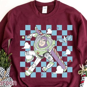 May include: A maroon sweatshirt with a checkered pattern and a cartoon image of Buzz Lightyear from Toy Story. Buzz is wearing his space ranger suit and is flying in his spaceship. The text "To Infinity and Beyond" is printed on the spaceship.