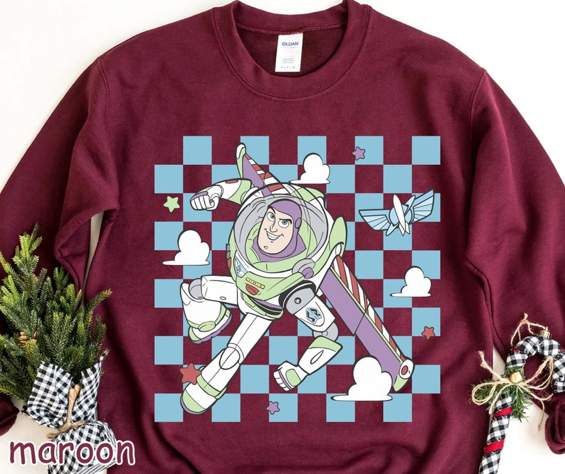 May include: A maroon sweatshirt with a checkered pattern and a cartoon image of Buzz Lightyear from Toy Story. Buzz is wearing his space ranger suit and is flying in his spaceship. The text "To Infinity and Beyond" is printed on the spaceship.