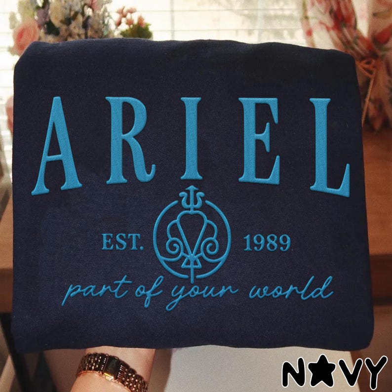 May include: Navy blue sweatshirt with the name "ARIEL" embroidered in teal. Below the name is a teal emblem with the text "EST. 1989". The phrase "part of your world" is embroidered in teal. The word "NAVY" is in the bottom right corner.