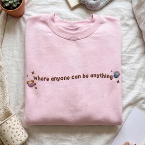K&ouml;nnte beinhalten: Ein hellrosa Sweatshirt mit Rundhalsausschnitt und dem gestickten braunen Schriftzug "where anyone can be anything" auf der Brust. Das Design enth&auml;lt kleine gestickte Sternakzente und zwei bunte, kreisf&ouml;rmige Charakter-Icons an den Seiten.
