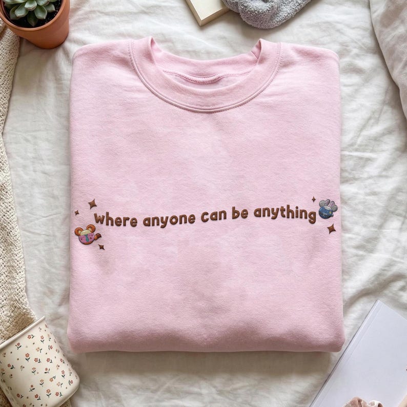 K&ouml;nnte beinhalten: Ein hellrosa Sweatshirt mit Rundhalsausschnitt und dem gestickten braunen Schriftzug "where anyone can be anything" auf der Brust. Das Design enth&auml;lt kleine gestickte Sternakzente und zwei bunte, kreisf&ouml;rmige Charakter-Icons an den Seiten.