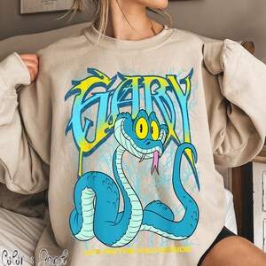 Op de afbeelding: Een zandkleurige sweatshirt met een cartoon slangenontwerp. Het woord "GARY" is geschreven in blauwe en gele graffiti-letters. De slang is blauw met gele ogen en een roze tong. De tekst "LIFE ON THE WILD SIDE" staat eronder.