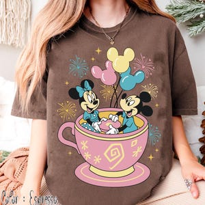 May include: Brown t-shirt with a cartoon of Mickey and Minnie Mouse in a pink teacup, balloons, and fireworks. The text "Color: Espresso" is at the bottom left of the image.