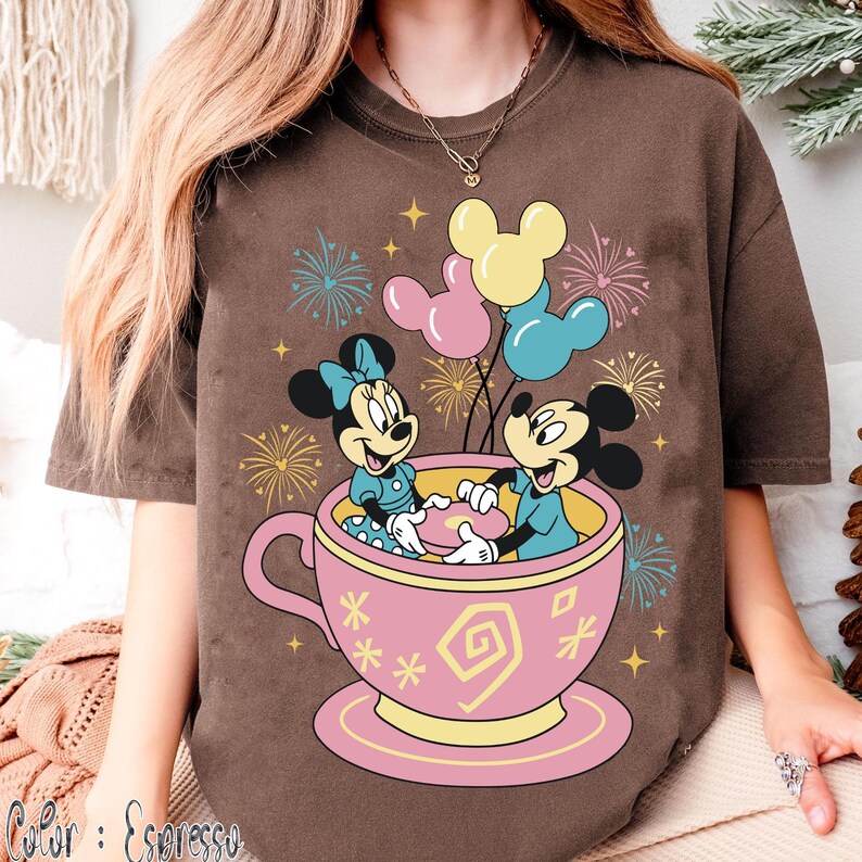 May include: Brown t-shirt with a cartoon of Mickey and Minnie Mouse in a pink teacup, balloons, and fireworks. The text "Color: Espresso" is at the bottom left of the image.