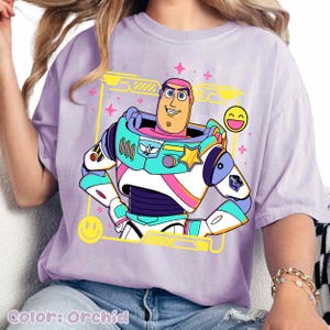 May include: Orchid-colored t-shirt featuring a colorful graphic of a cartoon character in a space suit. The design includes yellow, pink, blue, and white accents, with smiley faces and a yellow frame. The text "Color: Orchid" is at the bottom.