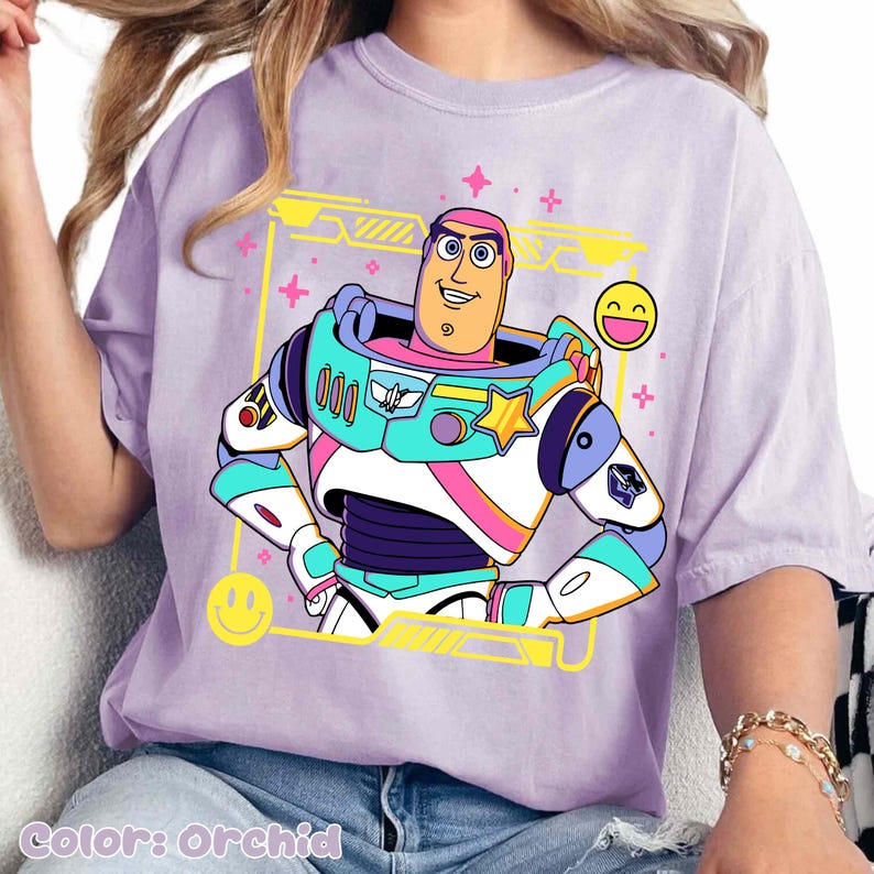 May include: Orchid-colored t-shirt featuring a colorful graphic of a cartoon character in a space suit. The design includes yellow, pink, blue, and white accents, with smiley faces and a yellow frame. The text "Color: Orchid" is at the bottom.