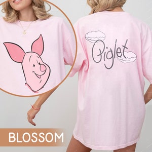 May include: A pink t-shirt with a Piglet cartoon character on the front and the word "Piglet" with clouds on the back. The shirt is labeled "BLOSSOM".