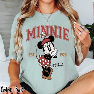 May include: A light green t-shirt featuring a graphic of Minnie Mouse. The design includes the word "MINNIE" in red block letters, the text "EST 1928", and Minnie Mouse wearing a red polka dot dress and hat. The word "Minnie" is also written in script.