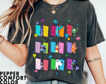 May include: A dark grey t-shirt with a colorful graphic print of characters from the movie Monsters Inc. The characters are peeking out of colorful doors with stars and flowers around them. The text "Mon Océan" is written on the right sleeve of the shirt.