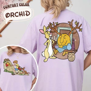May include: Orchid-colored t-shirt featuring a cartoon graphic of a rabbit and a smiling yellow character with antlers. The graphic includes the text "60 YEARS". The shirt also has a smaller graphic of a boy and a bear. The shirt is a Comfort Color.