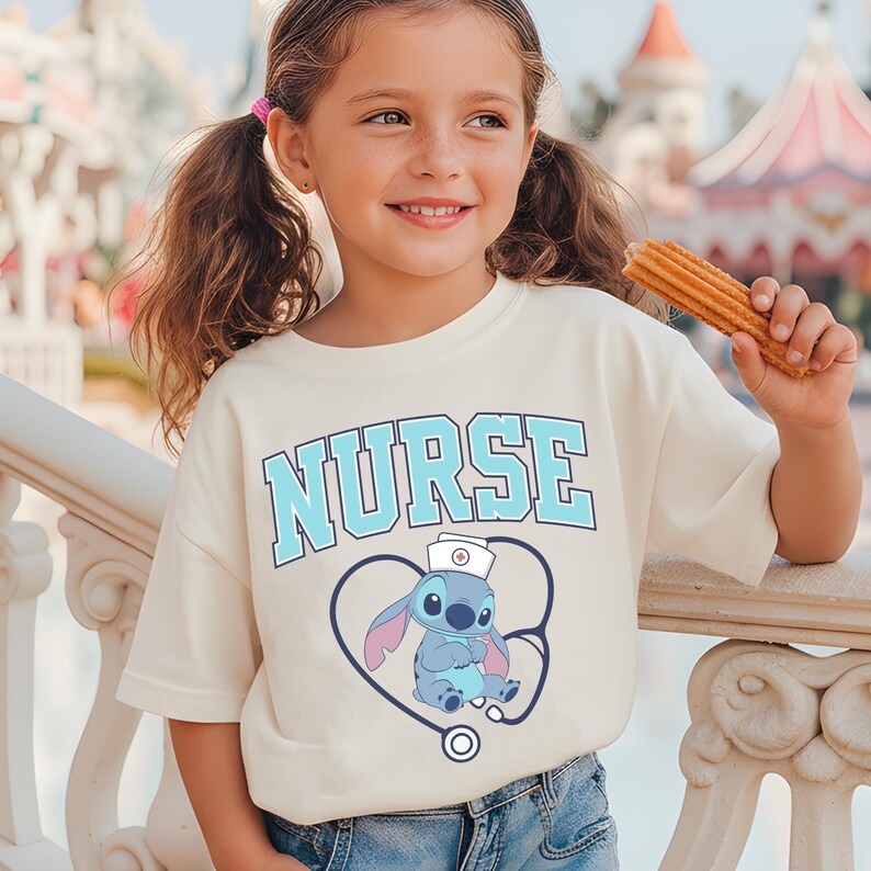 May include: A cream-colored t-shirt featuring the word "NURSE" in light blue block letters. Below the word is a graphic of a blue cartoon character wearing a nurse's hat, inside a heart-shaped stethoscope. The girl is holding a churro.