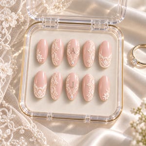 May include: A set of ten oval-shaped press-on nails in a soft blush pink hue, featuring intricate white lace-like relief patterns, small pearl accents, and the silver letters A and J centered on two of the nails, all displayed in a clear case.
