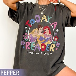 May include: Dark gray t-shirt featuring a colorful graphic of three cartoon princesses reading a book inside a circle. The text "TODAY A READER Tomorrow A Leader" is printed around the circle with floral accents. The shirt is a relaxed fit.