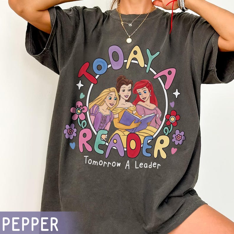 May include: Dark gray t-shirt featuring a colorful graphic of three cartoon princesses reading a book inside a circle. The text "TODAY A READER Tomorrow A Leader" is printed around the circle with floral accents. The shirt is a relaxed fit.