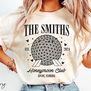 May include: A white t-shirt with a black graphic design featuring the text "THE SMITHS Honeymoon Club EST. 2024 EPCOT, FLORIDA" and a black outline of the Spaceship Earth attraction at Epcot.