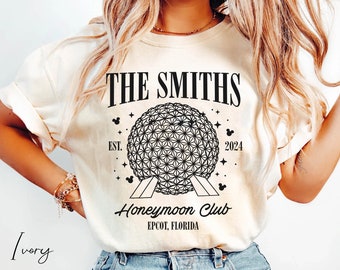 May include: A white t-shirt with a black graphic design featuring the text "THE SMITHS Honeymoon Club EST. 2024 EPCOT, FLORIDA" and a black outline of the Spaceship Earth attraction at Epcot.