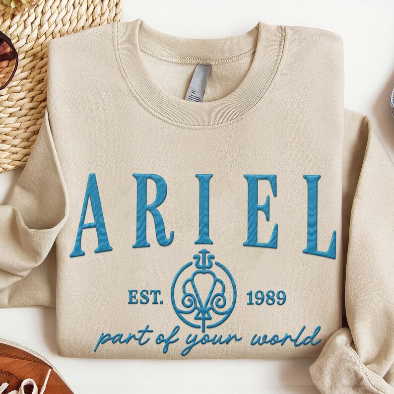 May include: Beige crewneck sweatshirt with the name "ARIEL" embroidered in blue. Below the name is a blue emblem with the text "EST. 1989" and "part of your world". The sweatshirt is made of a soft material.