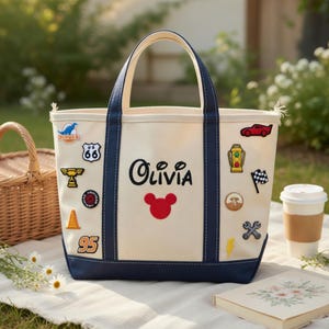 May include: Cream-colored tote bag with navy blue trim and handles. The bag features the name "Olivia" in black script, a red Mickey Mouse head, and various embroidered patches, including a race car and a Route 66 sign. A picnic basket and coffee cup are in the background.