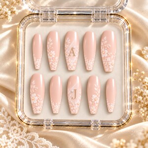May include: A set of ten press-on nails in a soft nude pink shade, featuring white floral lace patterns, small pearl accents, and the gold letters A and J on two of the nails. The nails are arranged in a clear plastic display case with a gold trim.