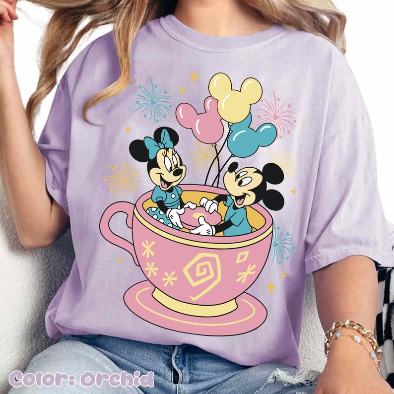 May include: Orchid-coloured t-shirt with a graphic of Mickey and Minnie Mouse in a teacup with balloons. The teacup is pink with yellow accents, and the characters are in blue outfits. Fireworks and stars are in the background.
