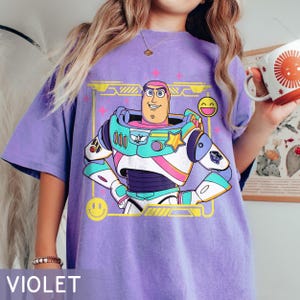 May include: Violet-colored t-shirt featuring a colorful graphic of Buzz Lightyear from Toy Story. The graphic includes a smiling Buzz, stars, and a yellow frame with smiley faces. The word "VIOLET" is printed at the bottom of the shirt.