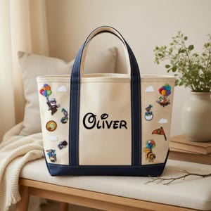 May include: A cream-colored canvas tote bag with dark navy blue handles and a reinforced base. The bag features the name Oliver printed in black script in the center, surrounded by various colorful embroidered patches of cartoon characters.