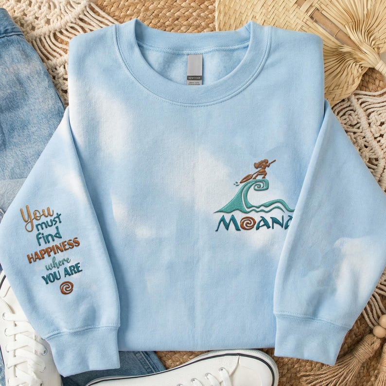 May include: A light blue tie-dye sweatshirt featuring an embroidered Moana graphic on the chest with a wave and a character on a boat. The left sleeve is embroidered with the text: You must find HAPPINESS where YOU ARE and a spiral symbol.