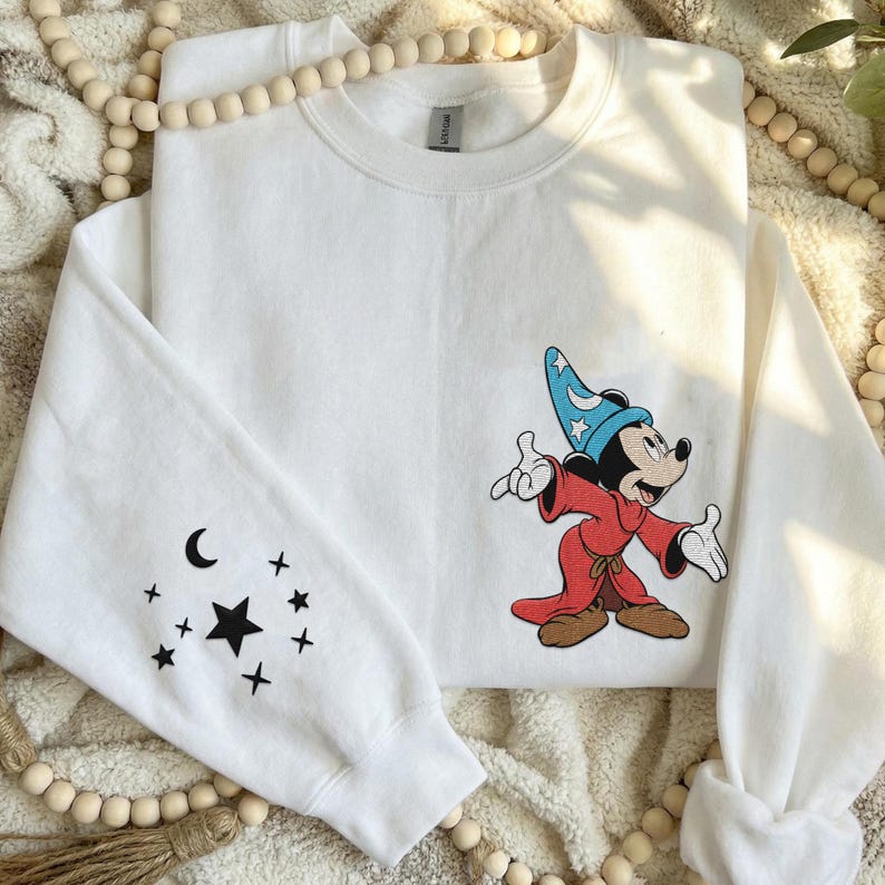 Puede incluir: Una sudadera blanca de cuello redondo con un gr&aacute;fico bordado de un rat&oacute;n de dibujos animados con un sombrero de mago azul y una t&uacute;nica roja en el pecho. La manga izquierda muestra un dise&ntilde;o negro impreso de una luna creciente y estrellas.