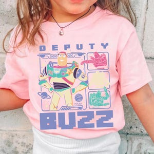 May include: A light pink t-shirt featuring a graphic of Buzz Lightyear from the Pixar movie Toy Story. The design includes the words "DEPUTY" and "BUZZ" in a pixelated font, along with images of Buzz Lightyear in various poses.