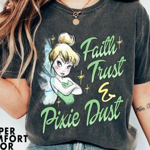 Może przedstawiać: Czarny T-shirt z grafiką w stylu vintage przedstawiającą Dzwoneczek z tekstem "Faith Trust & Pixie Dust" w kolorze zielonym i żółtym.