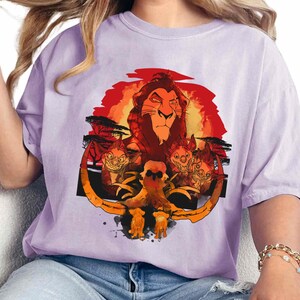 May include: A purple t-shirt with a graphic print of Simba, the lion from Disney's The Lion King, with a sunset background and hyenas in the foreground.