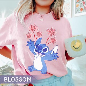 May include: Pink t-shirt featuring a blue cartoon character with a large head and big eyes, with red and white fireworks. The word "BLOSSOM" is printed at the bottom of the shirt.