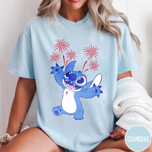 May include: Light blue t-shirt featuring a cartoon Stitch character with a happy expression, arms raised, and fireworks above. The design is in shades of blue and red. The shirt is a casual style.