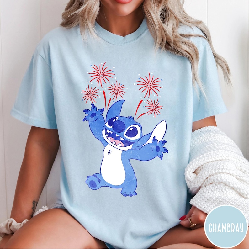 May include: Light blue t-shirt featuring a cartoon Stitch character with a happy expression, arms raised, and fireworks above. The design is in shades of blue and red. The shirt is a casual style.
