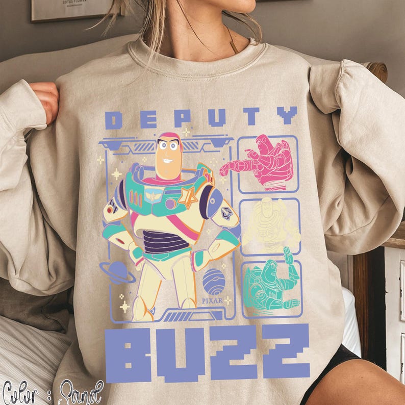 May include: Beige sweatshirt featuring a graphic of Buzz Lightyear with the text "DEPUTY BUZZ". The design includes images of Buzz in different poses and the Pixar logo. The sweatshirt has long sleeves.