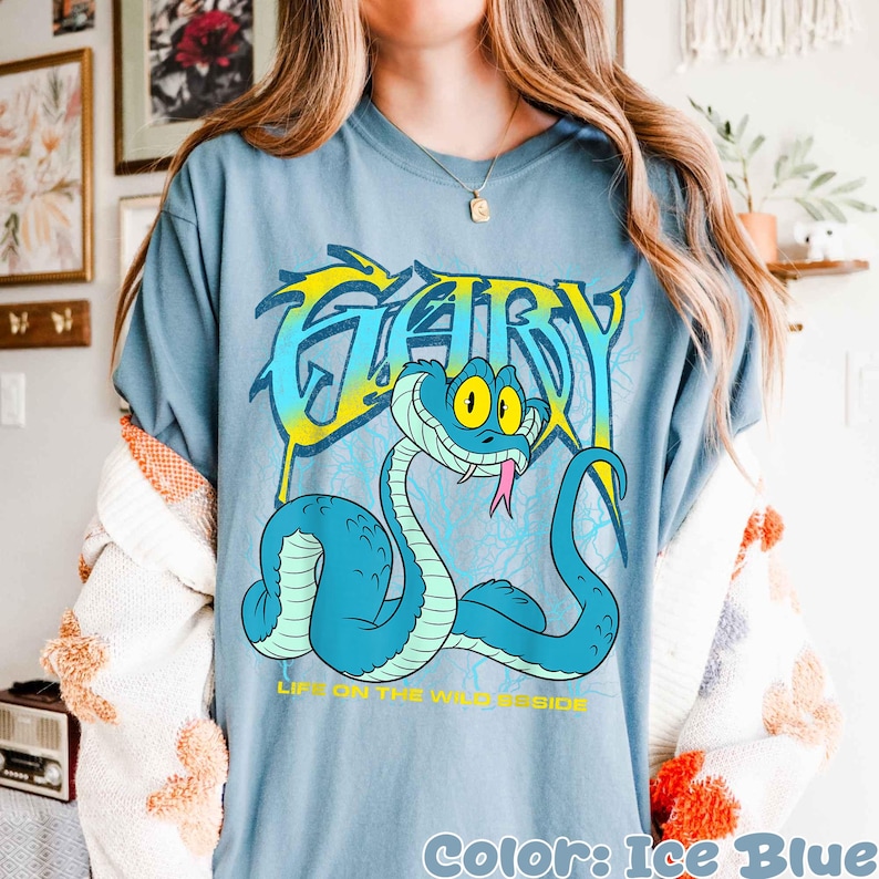 Op de afbeelding: Ijsblauw T-shirt met een cartoon slang en het woord "GARY" in een gestileerd lettertype. De slang is lichtblauw met gele accenten en een roze tong. De tekst "LIFE ON THE WILD SIDE" staat eronder.