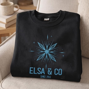 May include: A black crewneck sweatshirt featuring a light blue embroidered snowflake design centered on the chest. Below the snowflake, the text reads ELSA & CO in light blue capital letters, with the smaller text SINCE 2013 underneath.