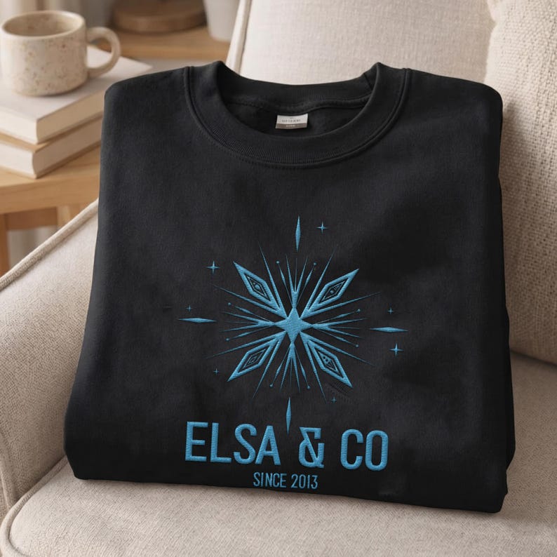 May include: A black crewneck sweatshirt featuring a light blue embroidered snowflake design centered on the chest. Below the snowflake, the text reads ELSA & CO in light blue capital letters, with the smaller text SINCE 2013 underneath.
