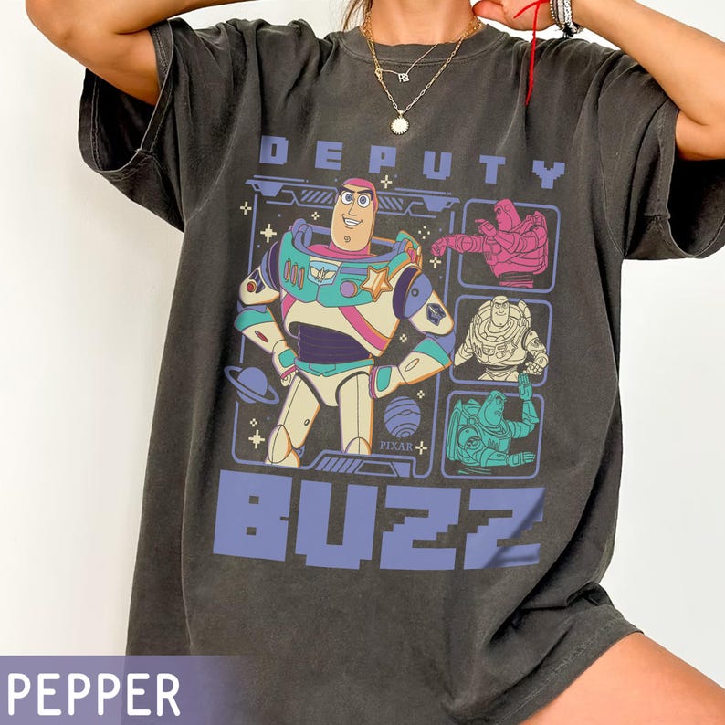 May include: Dark grey t-shirt featuring a graphic of Buzz Lightyear from Toy Story. The design includes the words "DEPUTY" and "BUZZ" in blue, along with multiple images of the character. The Pixar logo is also visible.