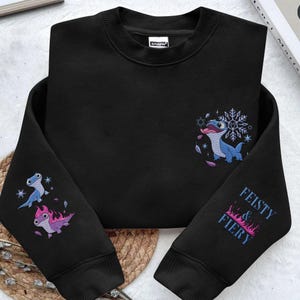 May include: A black long-sleeve crewneck sweatshirt featuring embroidered graphics. The chest displays a blue lizard with a snowflake. The left sleeve shows two lizards, one blue and one pink with flames. The right sleeve reads FEISTY & FIERY.