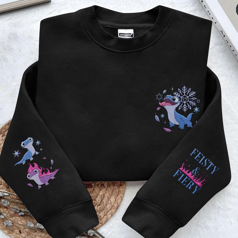 May include: A black long-sleeve crewneck sweatshirt featuring embroidered graphics. The chest displays a blue lizard with a snowflake. The left sleeve shows two lizards, one blue and one pink with flames. The right sleeve reads FEISTY & FIERY.