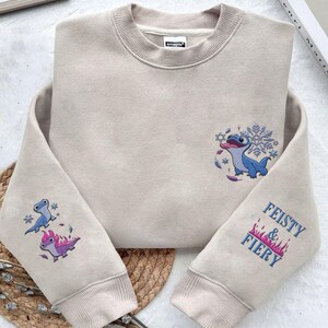 May include: A beige crewneck sweatshirt featuring embroidered designs. The chest displays a blue lizard-like creature with a snowflake. The left sleeve shows two small creatures, one blue and one purple with flames. The right sleeve reads FEISTY & FIERY.