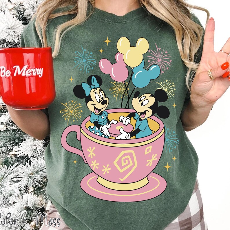 May include: Green t-shirt with a pink teacup design featuring Mickey and Minnie Mouse. Mickey and Minnie are inside the teacup, holding a heart. Mickey Mouse balloons float above. A red mug says "Be Merry".