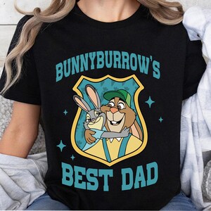 Puede incluir: Camiseta negra con un gr&aacute;fico que presenta dos conejos de dibujos animados abraz&aacute;ndose dentro de un escudo. El texto turquesa "BUNNYBURROW'S BEST DAD" est&aacute; impreso arriba y abajo de la imagen.