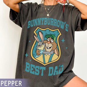 Puede incluir: Camiseta gris oscuro con un dibujo animado de dos conejos abraz&aacute;ndose y el texto turquesa "BUNNYBURROW'S BEST DAD". El gr&aacute;fico est&aacute; dentro de un escudo. La camiseta tiene un corte holgado.