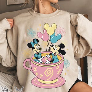 May include: Beige sweatshirt with a vibrant graphic of Mickey and Minnie Mouse in a pink teacup, holding balloons. The design includes fireworks and stars, creating a festive atmosphere. The sweatshirt is a comfortable, casual style.
