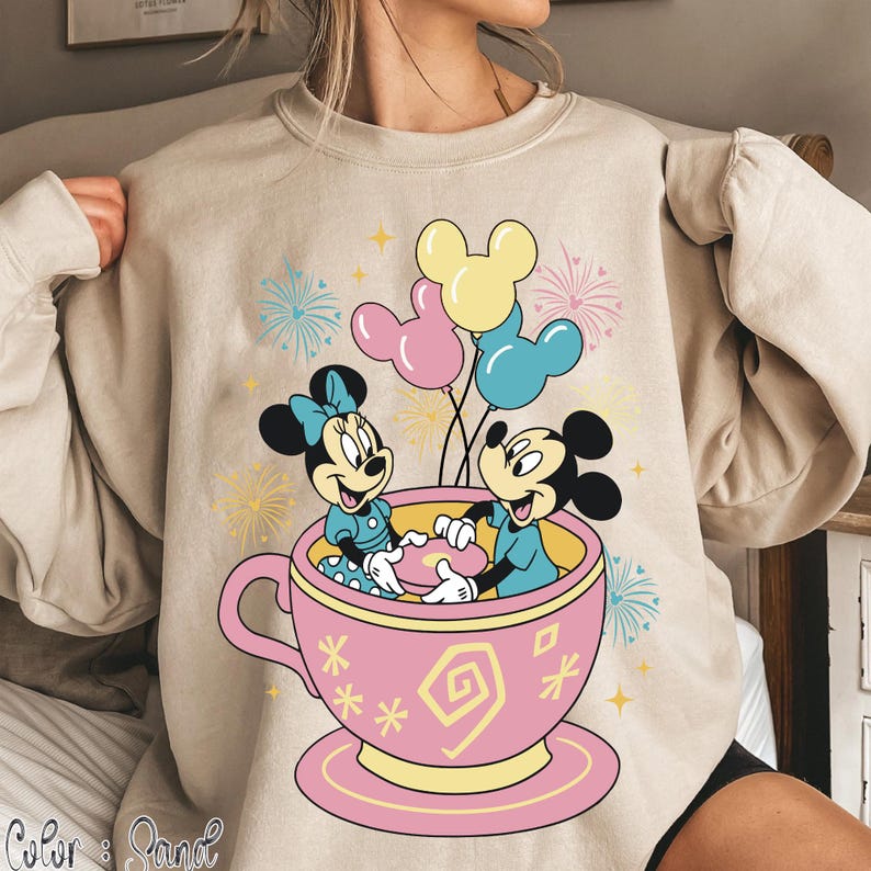 May include: Beige sweatshirt with a vibrant graphic of Mickey and Minnie Mouse in a pink teacup, holding balloons. The design includes fireworks and stars, creating a festive atmosphere. The sweatshirt is a comfortable, casual style.