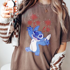 May include: Brown t-shirt featuring a blue cartoon character with fireworks. The character has large eyes and is smiling with its arms raised. Red and white fireworks are displayed above the character.