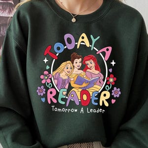 May include: Dark green sweatshirt with a colorful graphic featuring three cartoon princesses reading a book. The text "TODAY READER Tomorrow A Leader" is displayed in a circular design with floral accents.